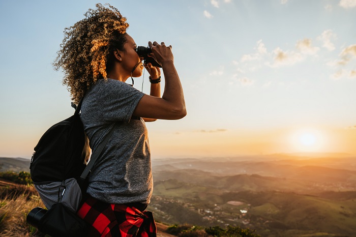 Woman hiking on top of mountain holding binoculars and looking out over the view as the sun sets