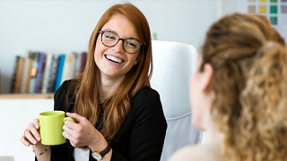 Une femme à lunettes, tenant une tasse vert citron, sourit à une autre femme.